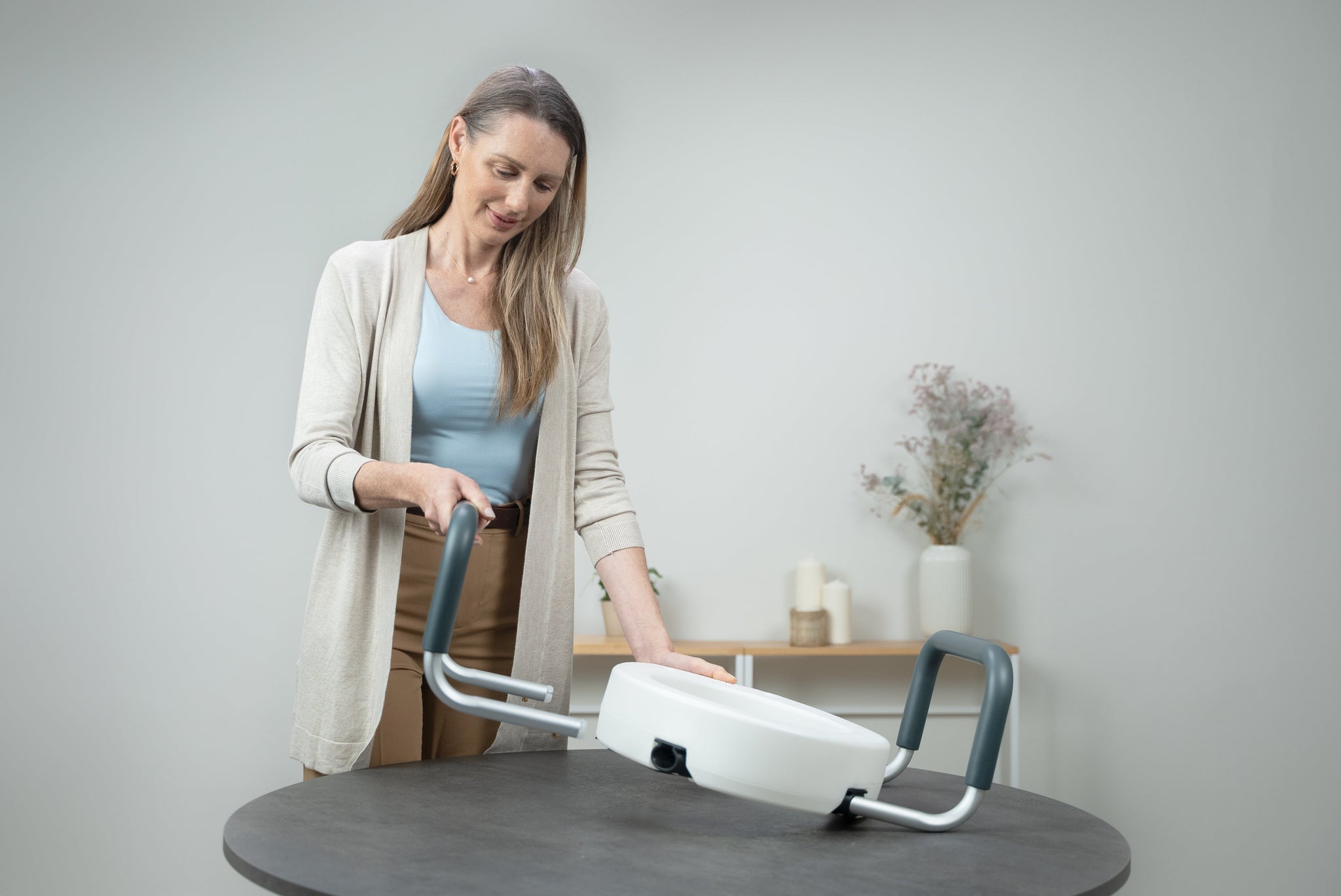 Woman demonstrating a raised toilet seat with padded handles, showing the elevated seat design and supportive armrests for safer sitting and standing in the bathroom.