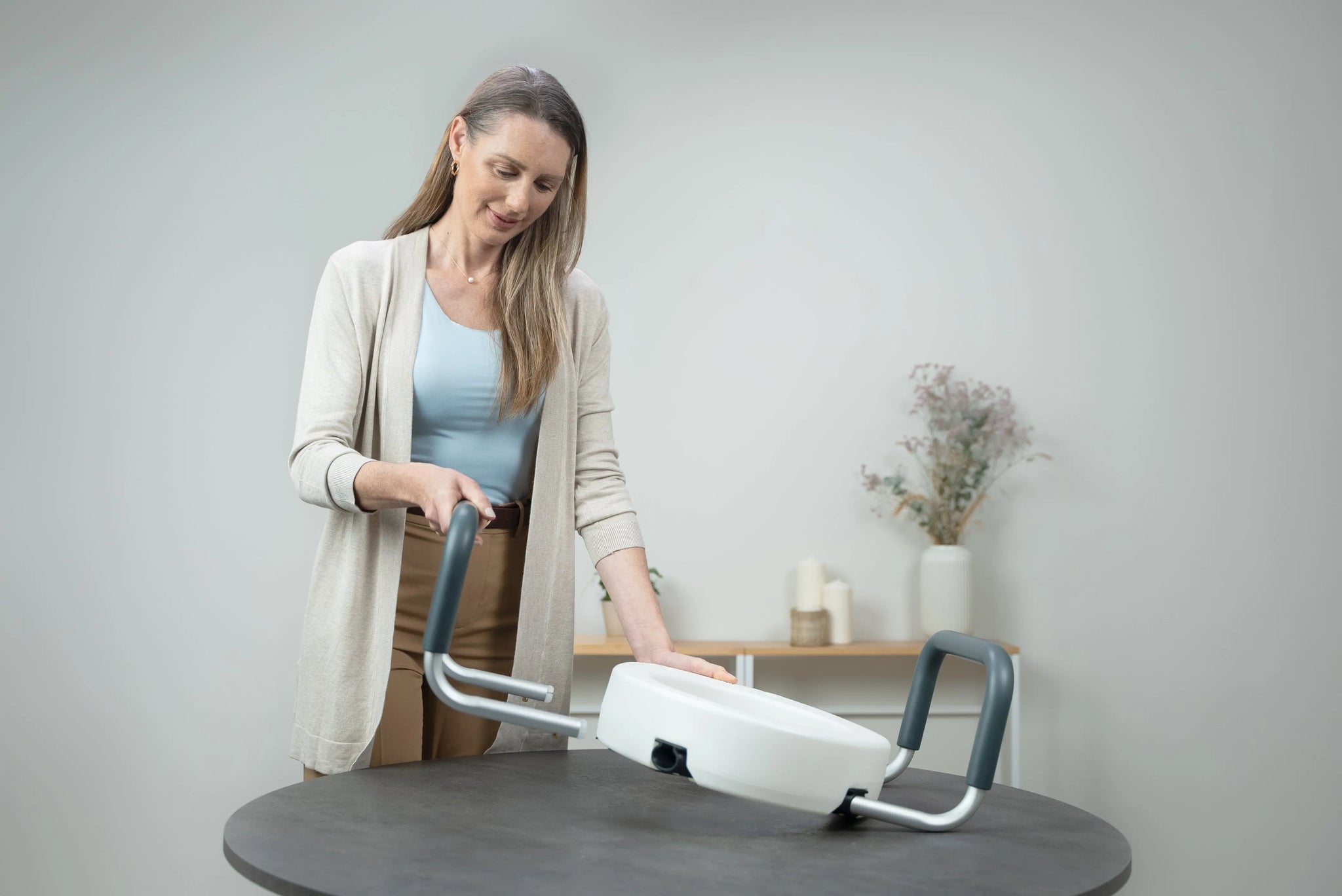 Woman demonstrating a raised toilet seat with padded handles, showing the elevated seat design and supportive armrests for safer sitting and standing in the bathroom.