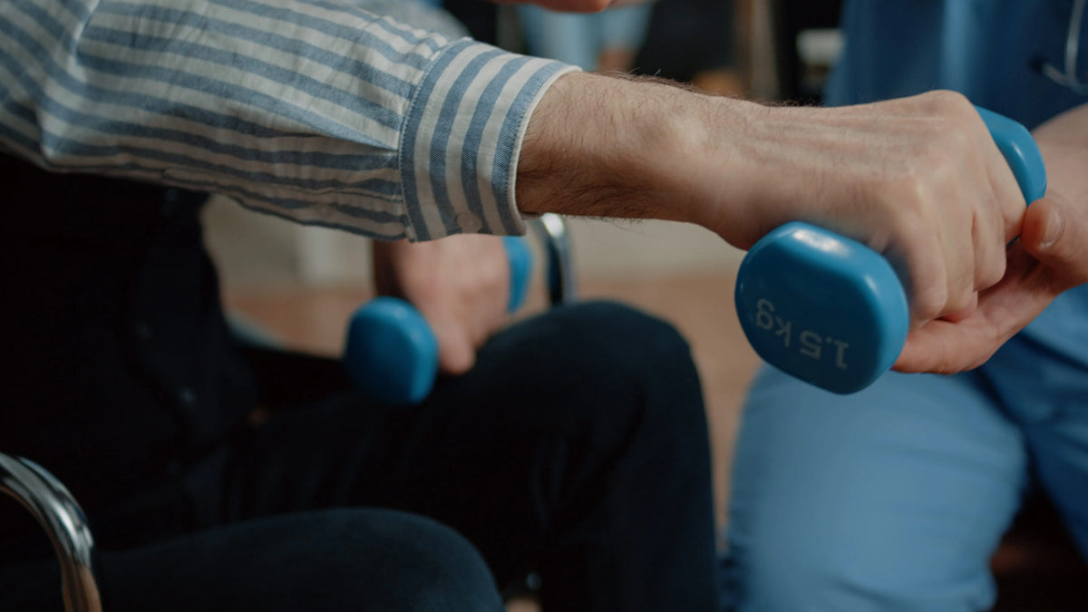 Older adult lifting blue dumbbells with a therapist supporting one hand during seated strength exercises.