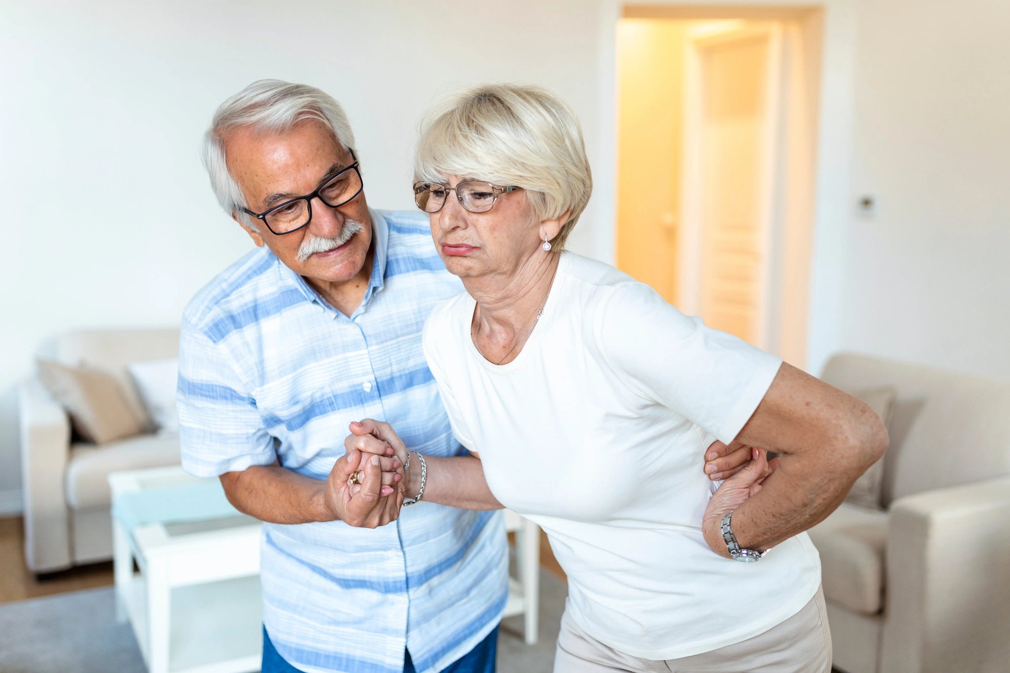 Senior woman experiencing sciatica pain while her husband supports her at home.