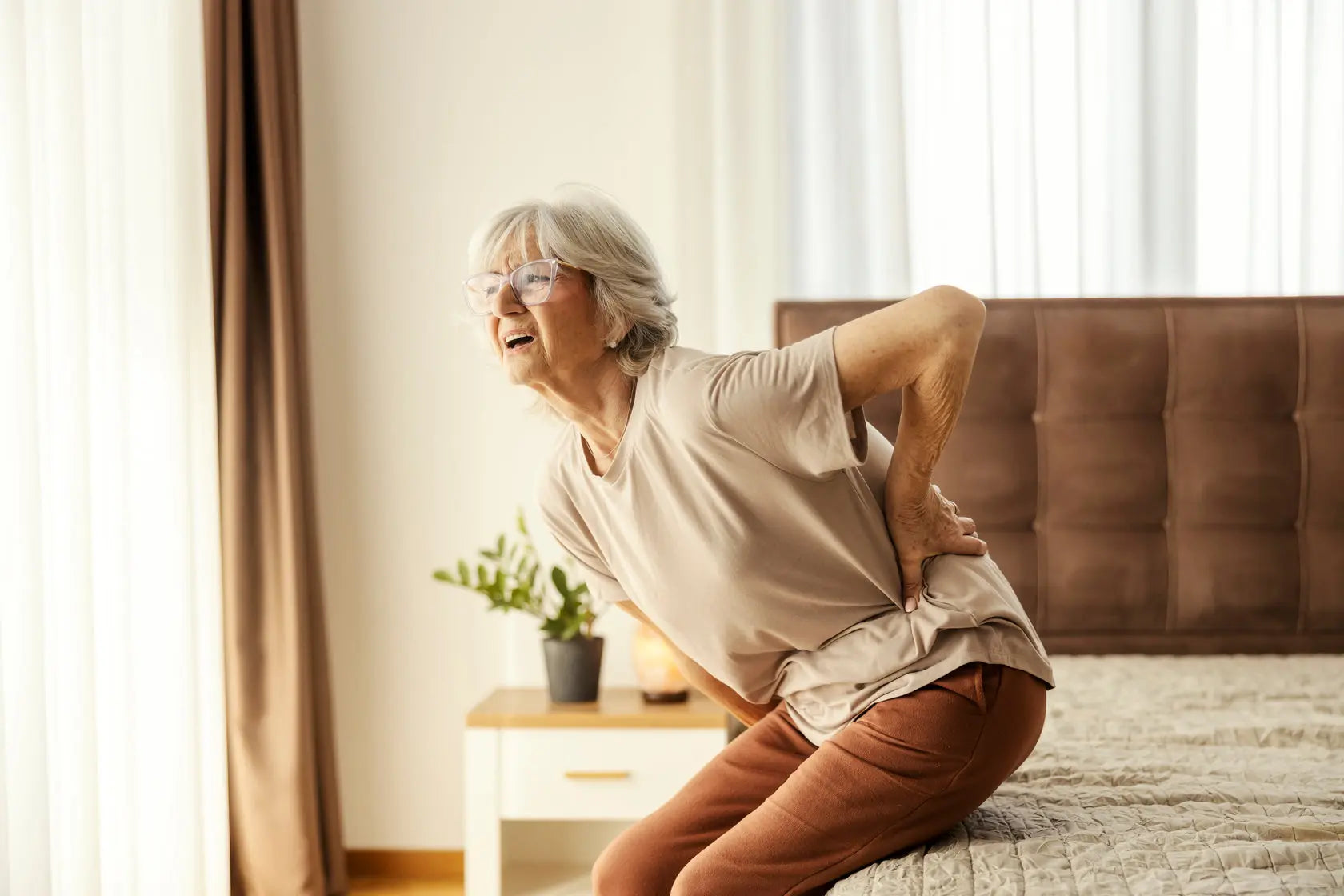 Older woman holding her lower back in pain while getting up from bed due to sciatica symptoms.