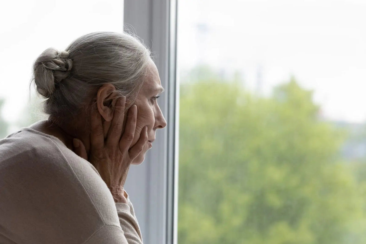 Older woman resting her face in her hands while looking out a window.