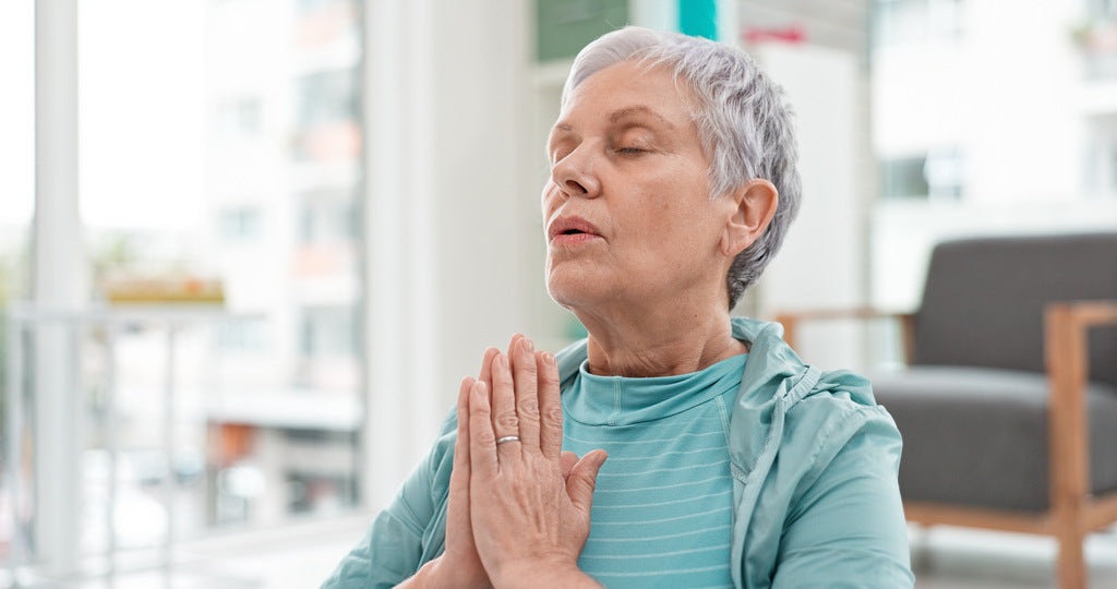 Senior woman practicing mindful breathing exercises at home with hands together and eyes closed.
